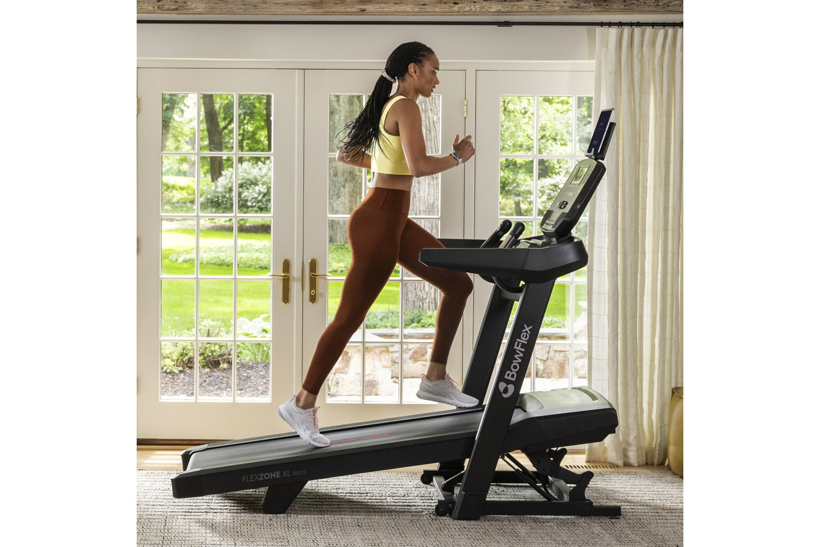 Woman running on Bowflex T9 treadmill wearing yellow sports bra and brown leggings in bright home gym with windows