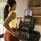 Woman wearing yellow sports bra and orange leggings running on BowFlex treadmill with tablet display showing fitness class
