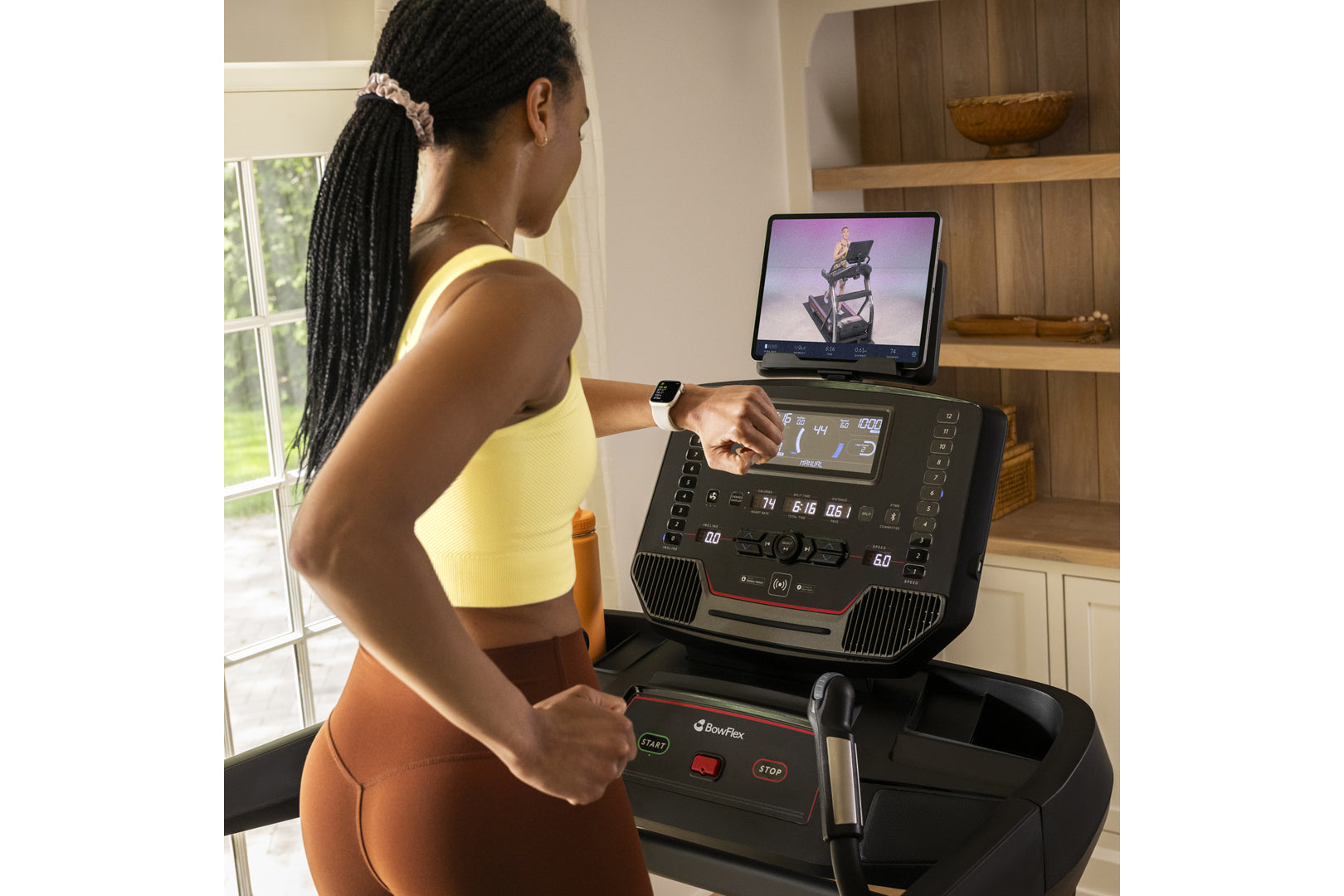 Woman wearing yellow sports bra and orange leggings running on BowFlex treadmill with tablet display showing fitness class