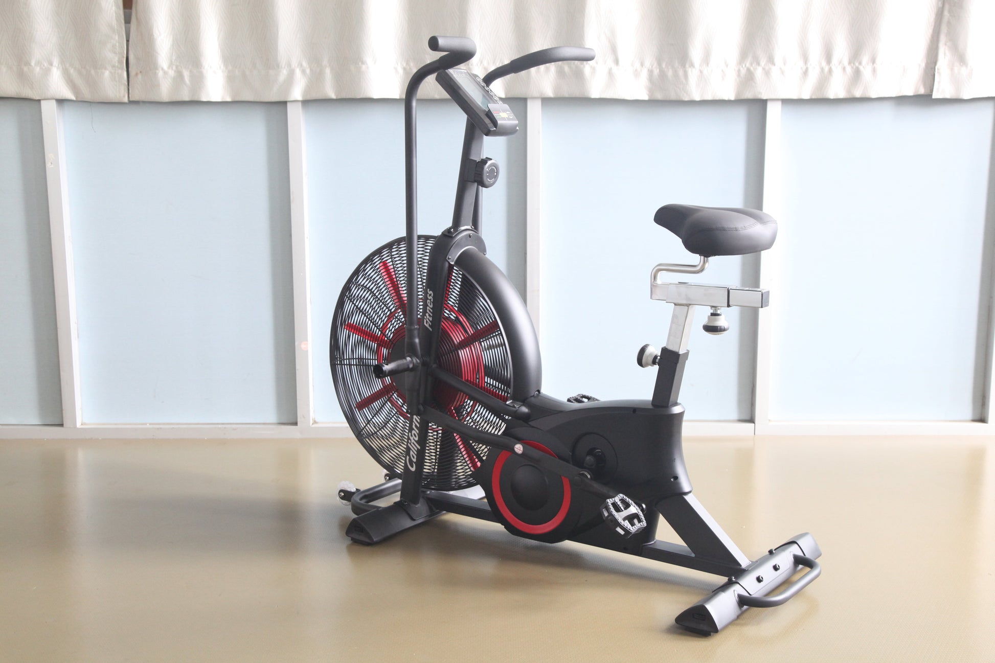 Black and red stationary air exercise bike with large fan wheel, curved handlebars, and padded seat on wood floor