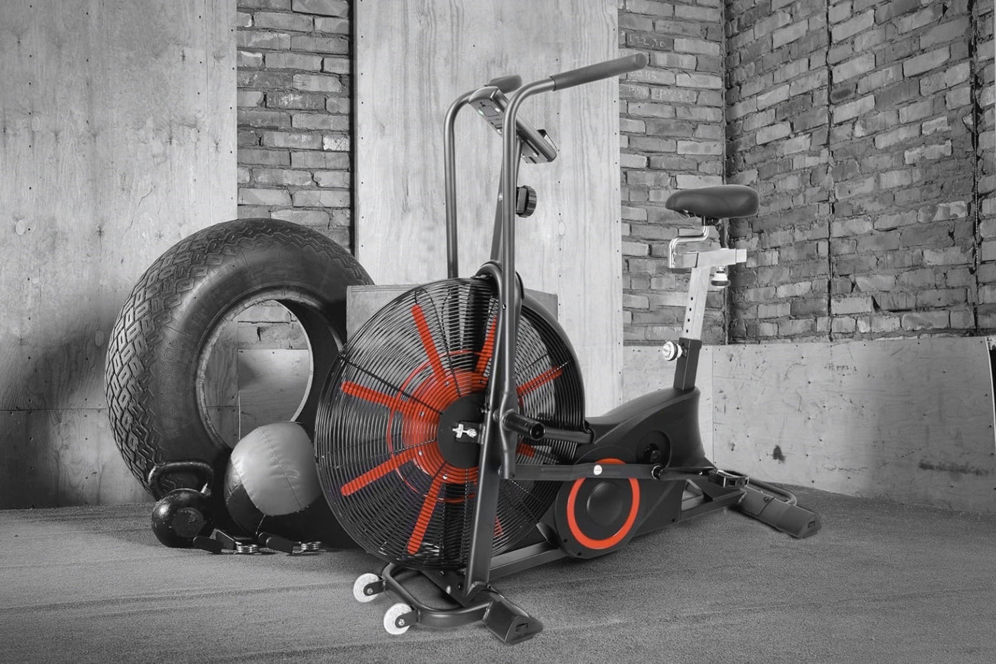 Black and red air exercise bike with large fan wheel, seat, and handlebars in industrial gym setting with tire and equipment