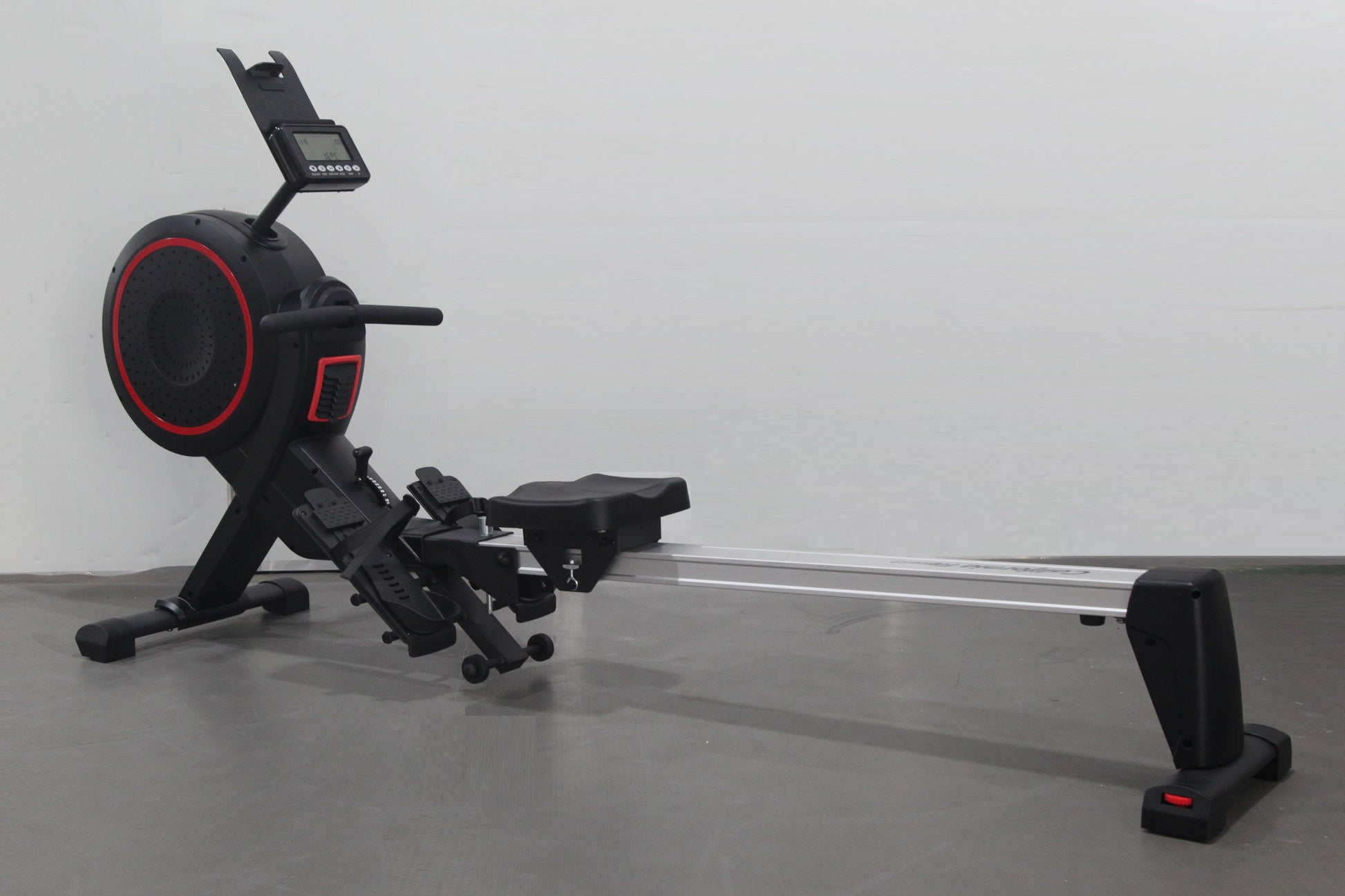 Black and red rowing machine with digital display monitor, foot pedals, and sliding seat on metal rail, on gray background