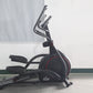 Black elliptical machine with red accents and dual handlebars, displayed on concrete floor against white wall