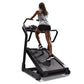 Woman using black incline trainer treadmill with handles, wearing gray tank top and black shorts on white background