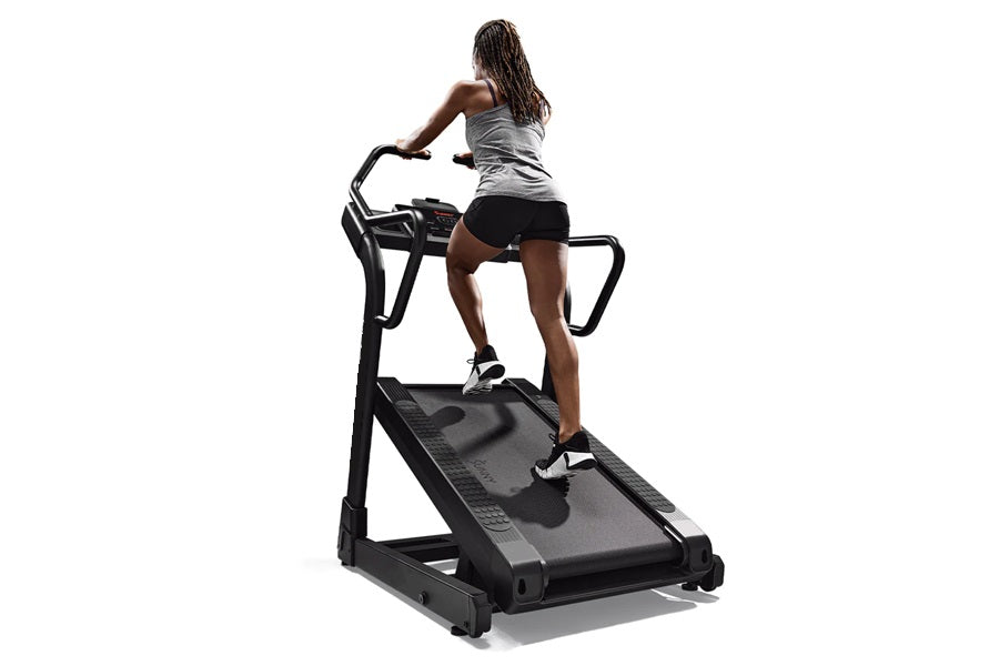 Woman using black incline trainer treadmill with handles, wearing gray tank top and black shorts on white background