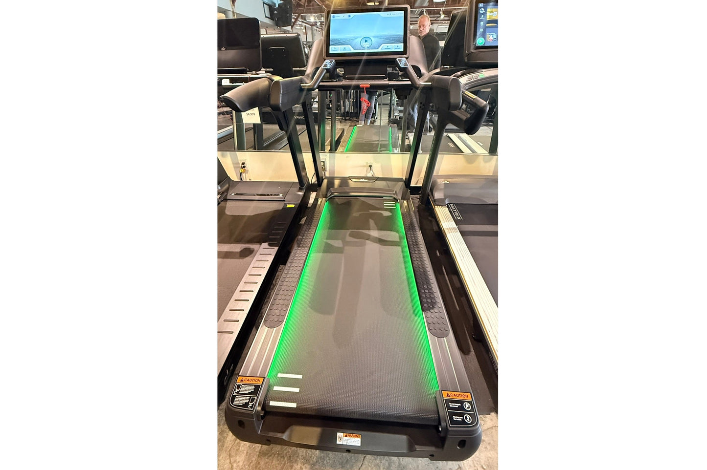 Black motorized treadmill with green-lit running deck, dual handles, and digital touchscreen console displaying workout inter