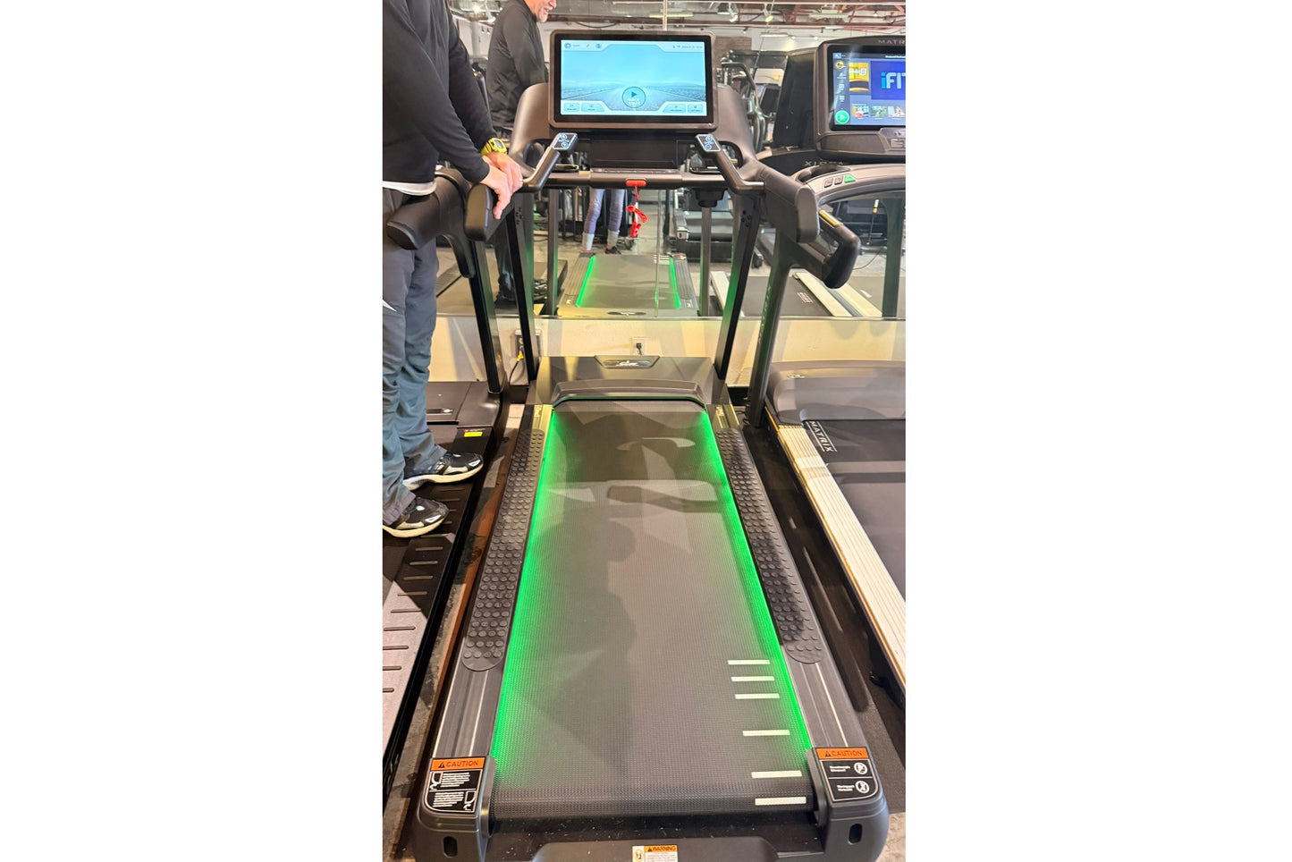 Black and green interactive treadmill with touch screen console, displayed at fitness showroom with person standing beside it