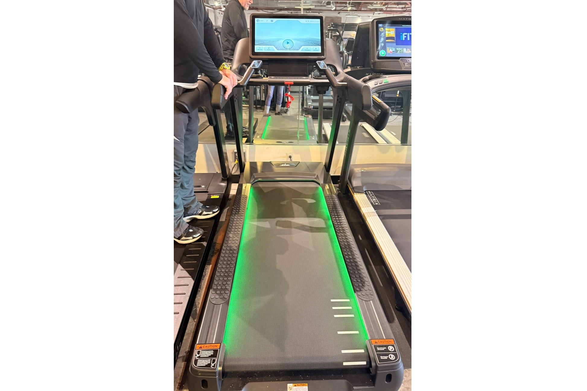Black and green interactive treadmill with touch screen console, displayed at fitness showroom with person standing beside it