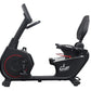 Black recumbent exercise bike with red accent ring on wheel, padded seat, adjustable armrests, and digital console display on