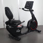 Black California Fitness recumbent exercise bike with digital display console, padded seat, and red accent wheel on dark floo