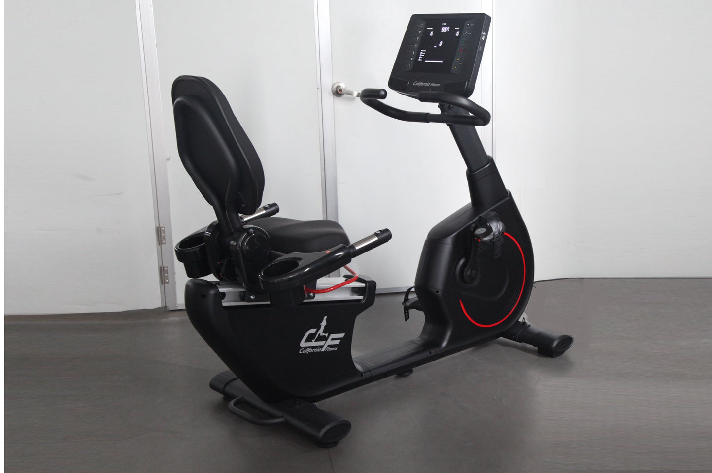 Black California Fitness recumbent exercise bike with digital display console, padded seat, and red accent wheel on dark floo