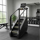Black and gray stair climber with digital display console in gym setting with dumbbells and equipment visible