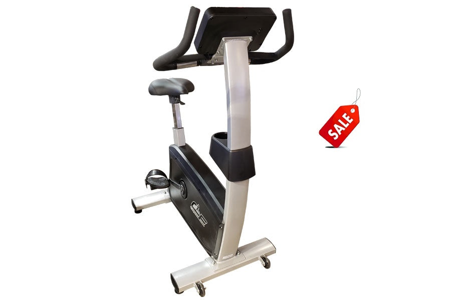 Upright exercise bike with black and silver frame, padded seat and handlebars, digital console display, and wheeled base on w