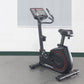 Black upright exercise bike with red accents, digital display console, and black padded seat on gray floor