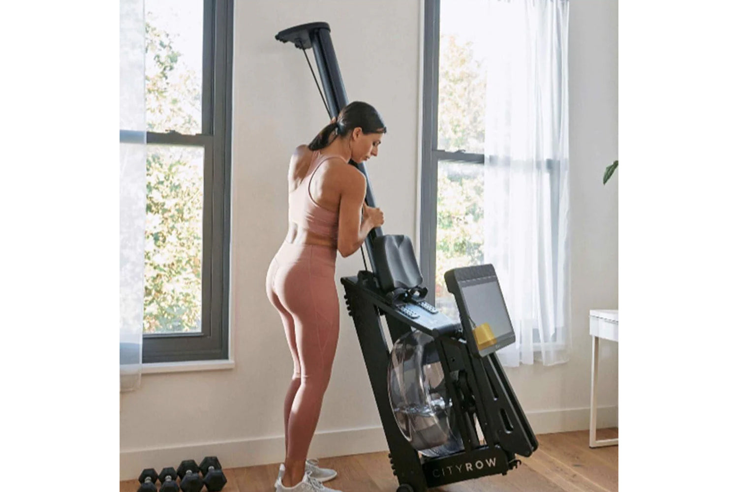 Woman wearing rose-pink ribbed sports bra and matching leggings using black CityRow rowing machine in bright home gym with la