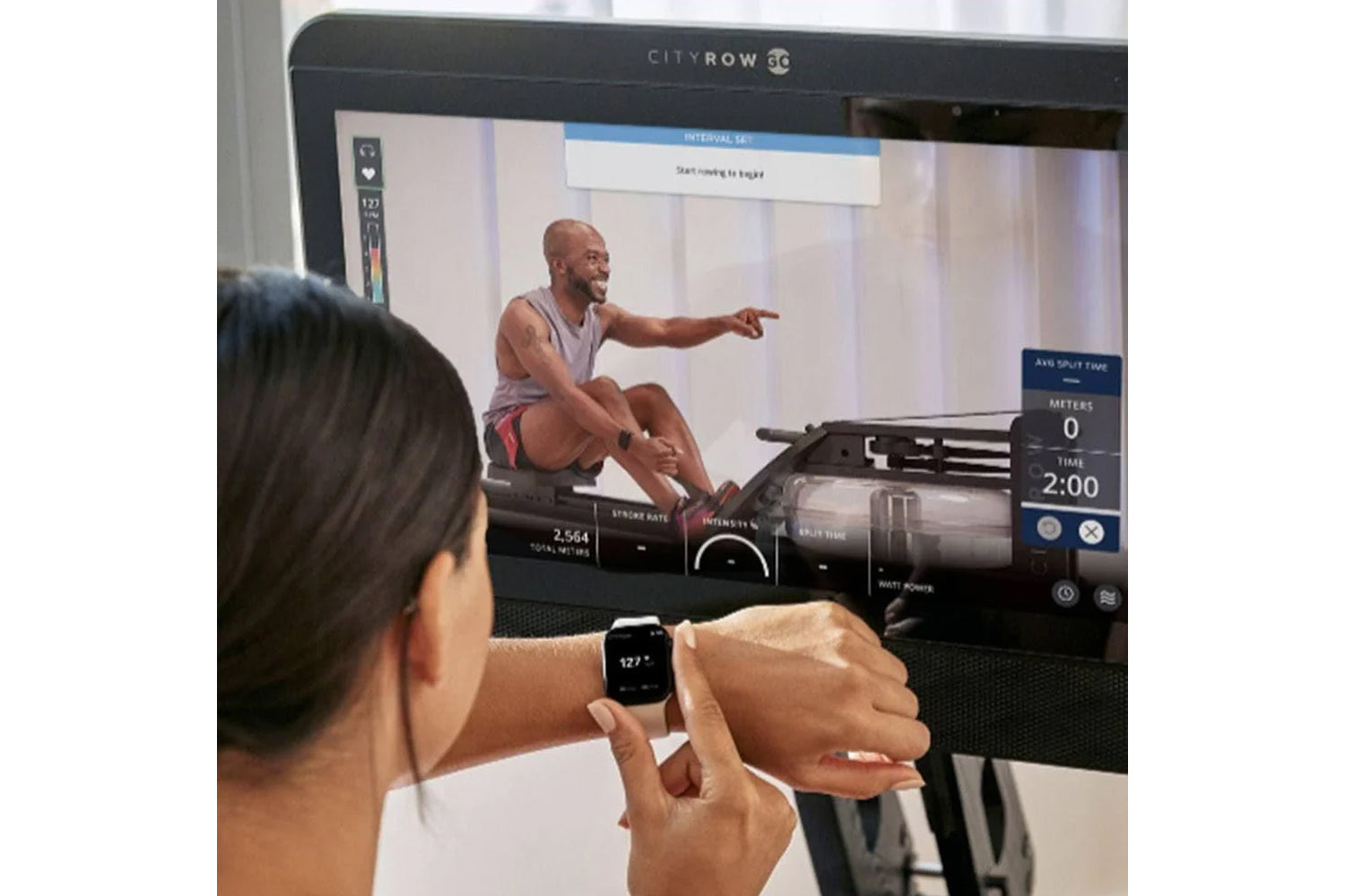 Person wearing smartwatch viewing CityRow GC rowing machine display screen showing instructor-led workout with performance me