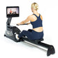 Woman in navy sports bra and leggings using black rowing machine with touchscreen display showing fitness class, on white bac