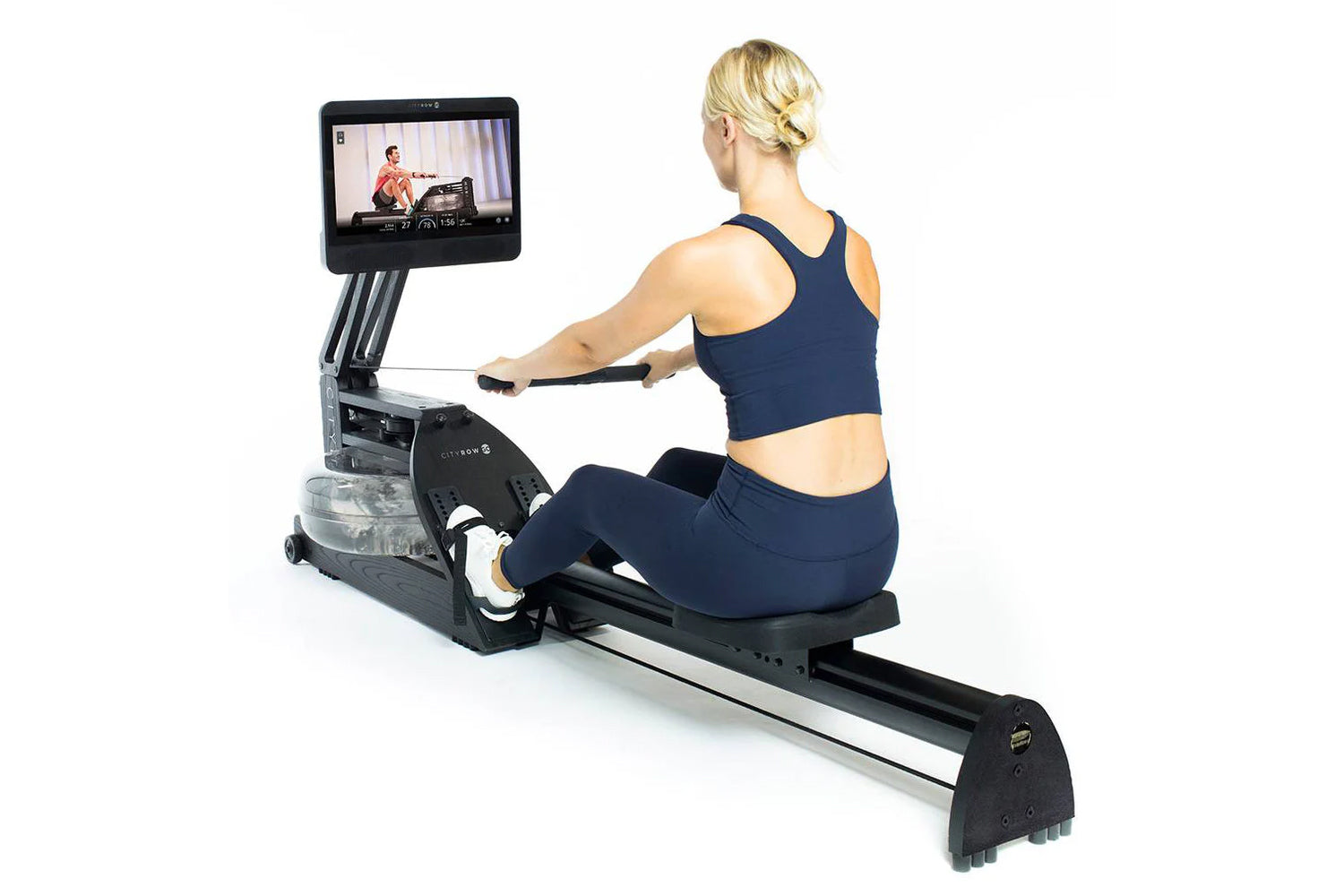 Woman in navy sports bra and leggings using black rowing machine with touchscreen display showing fitness class, on white bac