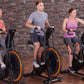 Three people on stationary exercise bikes with black frames and orange wheels against brick wall background