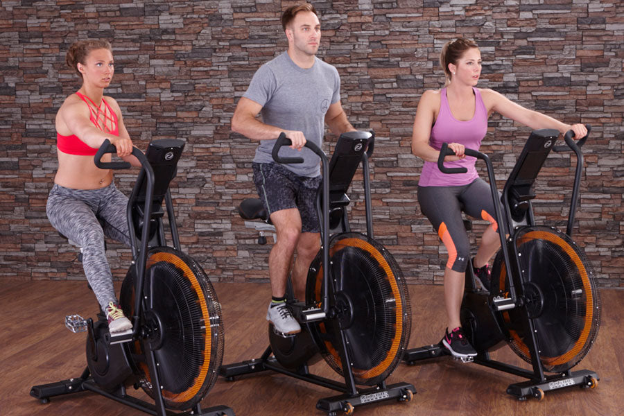Three people on stationary exercise bikes with black frames and orange wheels against brick wall background