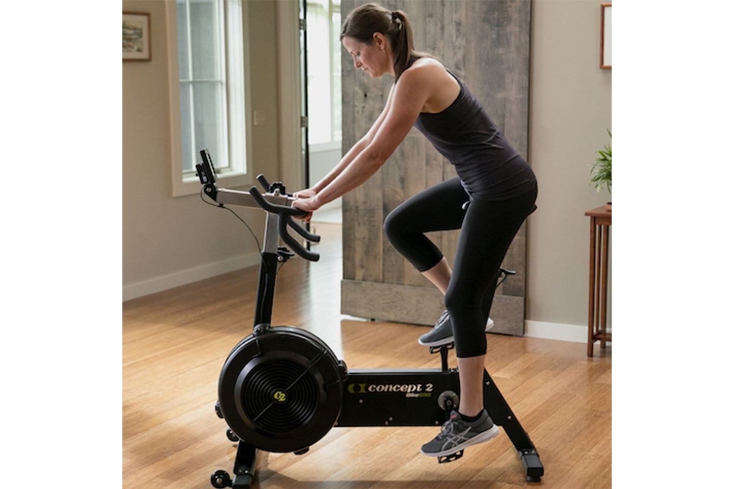 Woman wearing black tank top and capri leggings riding Concept2 BikeErg stationary bike in home gym setting