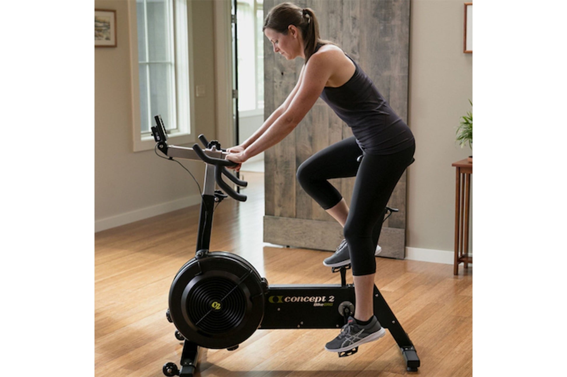 Woman wearing black tank top and capri leggings riding Concept2 BikeErg stationary bike in home gym setting