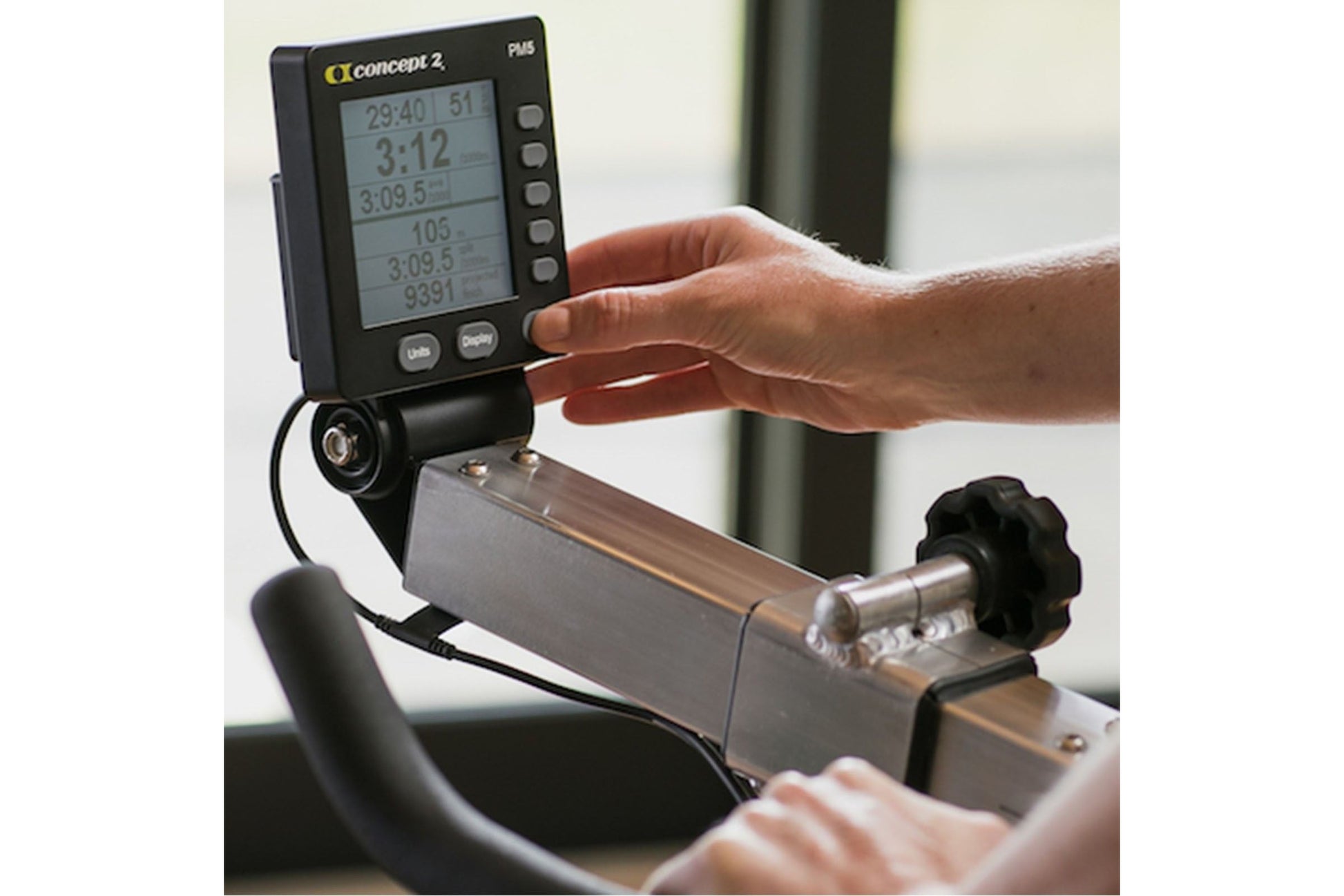 Concept2 PM5 black digital monitor displaying workout metrics, held by model's hand above stationary bike equipment