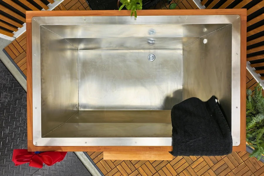 Stainless steel cold plunge tub with wooden frame and white surround, viewed from above on wooden deck with plants and red cl