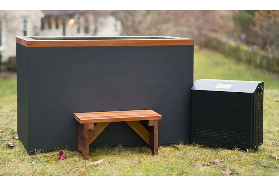 Dark grey cold plunge tub with copper trim and white interior, paired with wooden bench and black filtration unit on grass