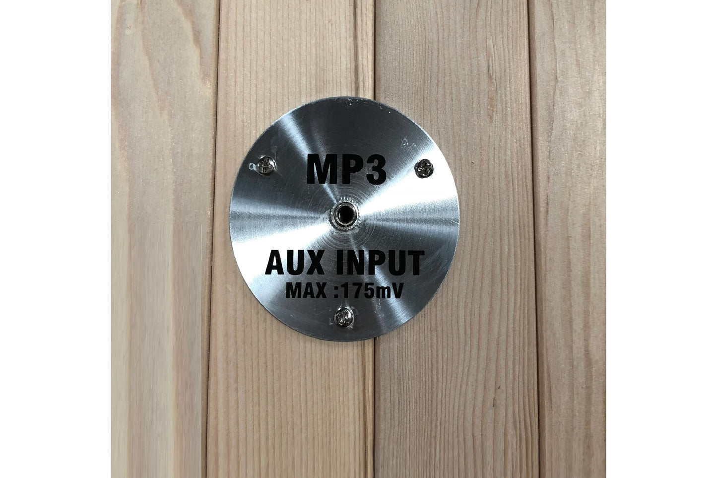 Brushed metal circular control panel with MP3 and AUX INPUT MAX 175mV text, mounted on light wood sauna wall
