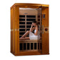 Two-person infrared sauna with golden-brown wooden frame and glass door, woman in white towel sitting inside on wooden bench,