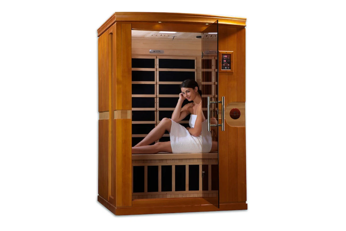 Two-person infrared sauna with golden-brown wooden frame and glass door, woman in white towel sitting inside on wooden bench,