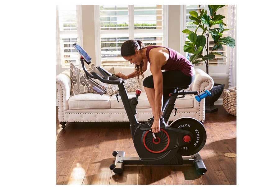 Woman in burgundy tank top using black Echelon Smart Connect stationary bike with red accent wheel in bright living room
