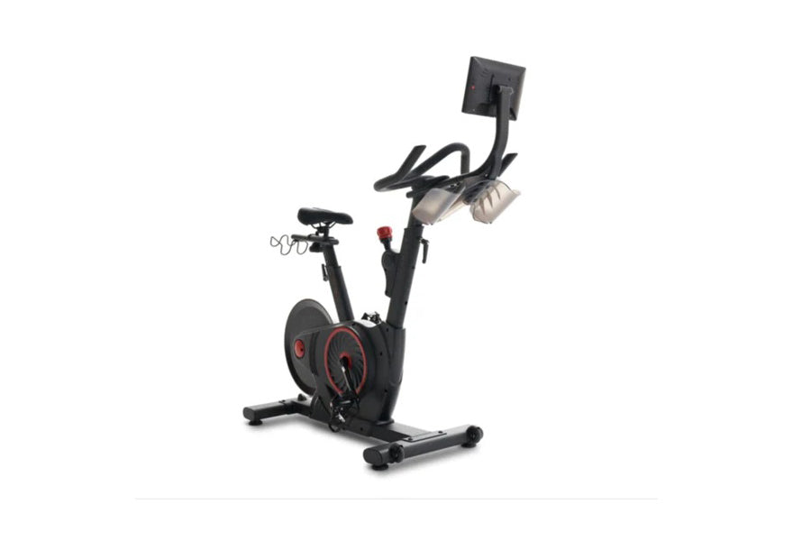 Black upright exercise bike with red accents, digital display mounted on adjustable arm, handlebars with controls, pedals, an