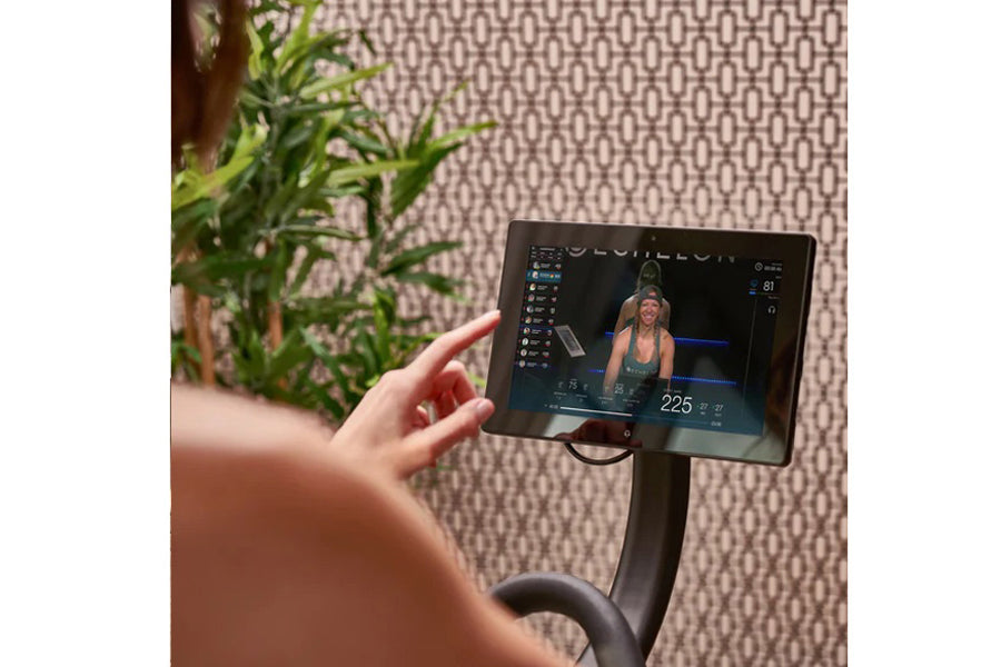 Person's hand on Echelon Smart Connect Bike EX-5s-10 with tablet displaying fitness class, potted plant visible