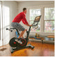 Man in red shirt and gray shorts riding black and red stationary exercise bike with digital screen in home gym