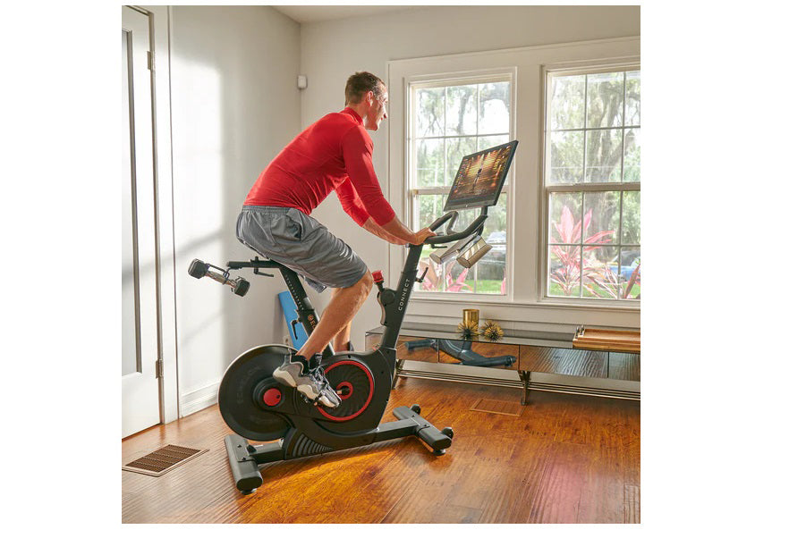 Man in red shirt and gray shorts riding black and red stationary exercise bike with digital screen in home gym