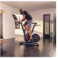 Man in navy shirt and gray shorts riding black stationary bike with red accents and tablet display in modern home gym setting