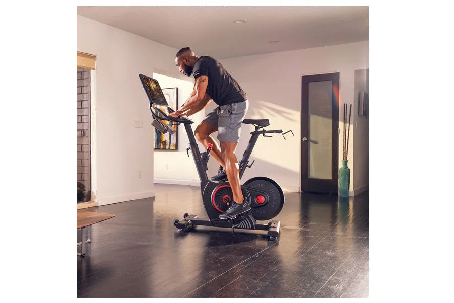 Man in navy shirt and gray shorts riding black stationary bike with red accents and tablet display in modern home gym setting