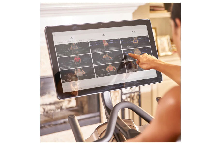 Person using touchscreen display on Echelon Smart Connect Bike EX-5s-22 showing workout class grid interface