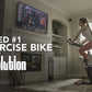 Woman in white tank top and burgundy leggings riding black and red stationary exercise bike in home gym with TV mounted on wa