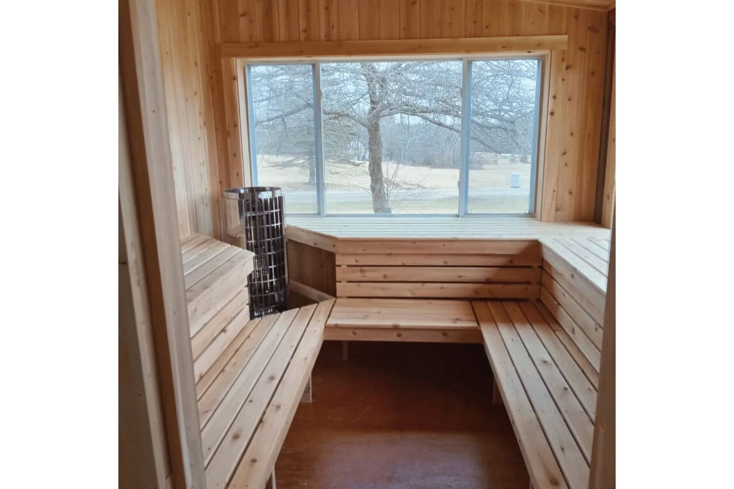 Interior of wooden sauna with light pine benches, metal heater basket, large windows overlooking winter landscape