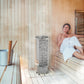 Woman wrapped in white towel relaxing on wooden sauna bench next to cylindrical Harvia electric sauna heater with built-in co