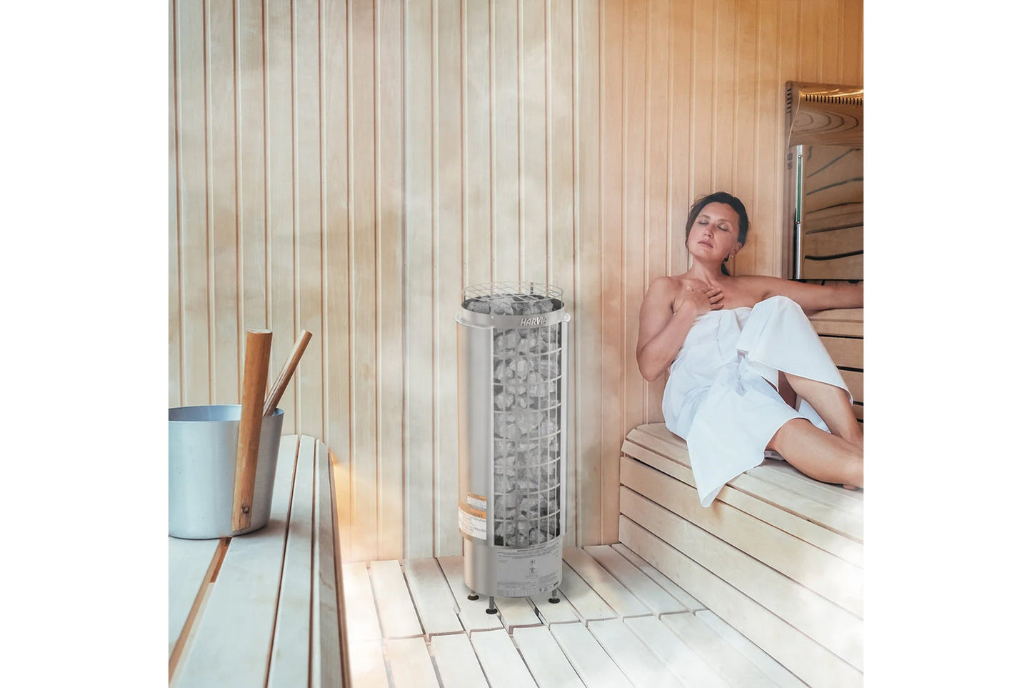 Woman wrapped in white towel relaxing on wooden sauna bench next to cylindrical Harvia electric sauna heater with built-in co