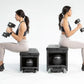 Two women performing dumbbell exercises on black FITBENCH STUDIO weight benches, wearing sports bras and leggings on white ba