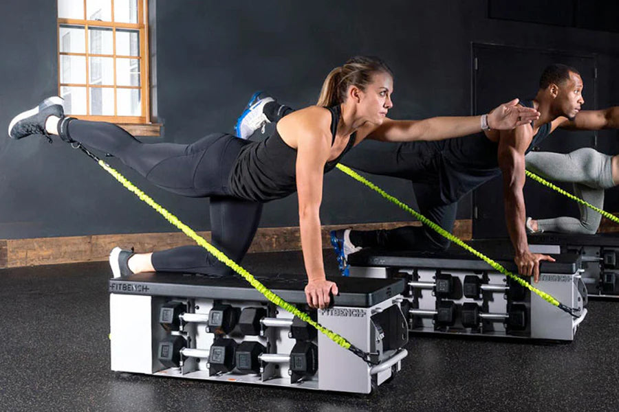 Two people performing plank exercises on FITBENCH STUDIO weight bench with neon yellow straps in gym setting