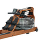 Brown wood and black rowing machine with digital monitor, water tank, and padded foot pedals on white background