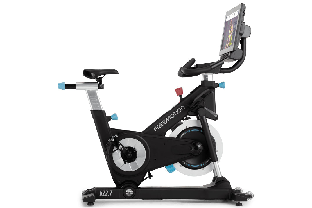 Freemotion CoachBike indoor cycle with black frame, white flywheel, blue accents, upright handlebars, and tablet holder displ