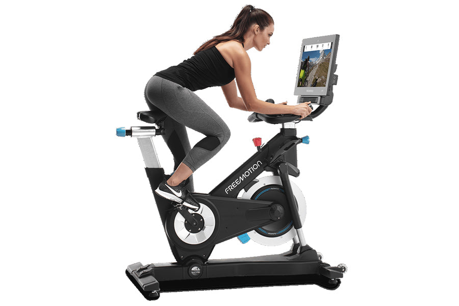 Woman exercising on black and white Freemotion CoachBike indoor cycle with tablet mounted on handlebars displaying fitness co
