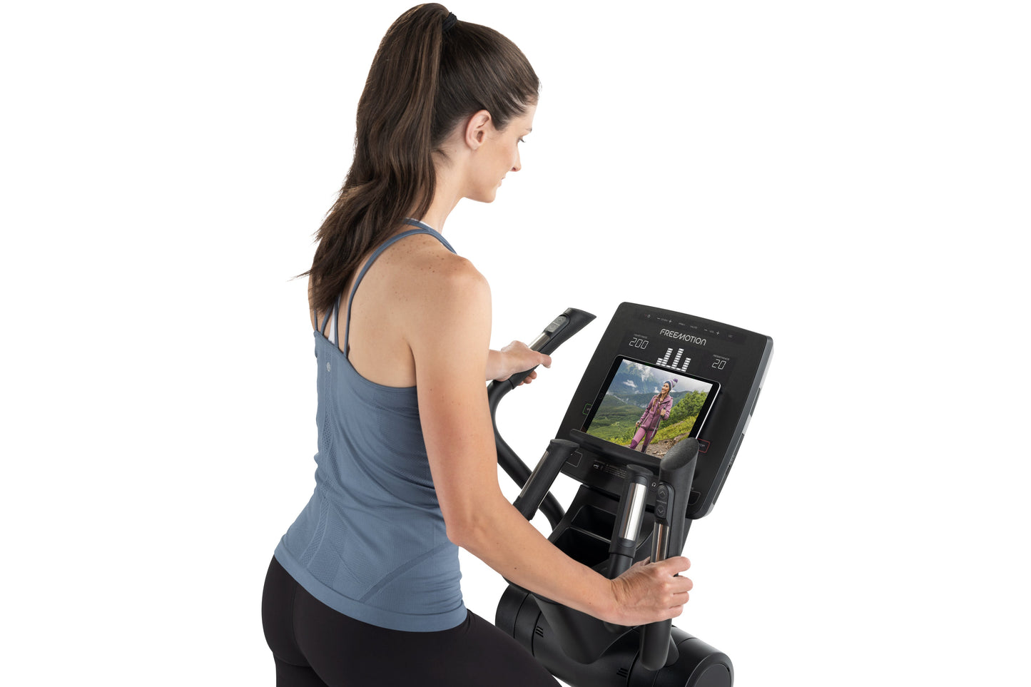 Woman in blue tank top using Freemotion e10.9b elliptical machine with display screen showing workout video, side view on whi
